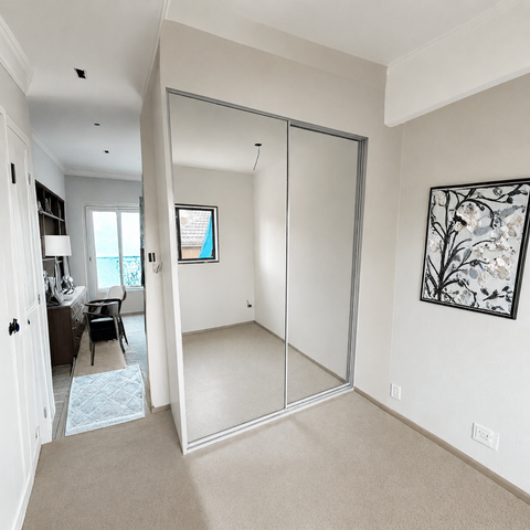 A modern bedroom featuring a large built-in wardrobe with two sliding glass doors framed in silver metal. The room has beige walls, beige carpet, and a framed black-and-white floral print on the right wall.