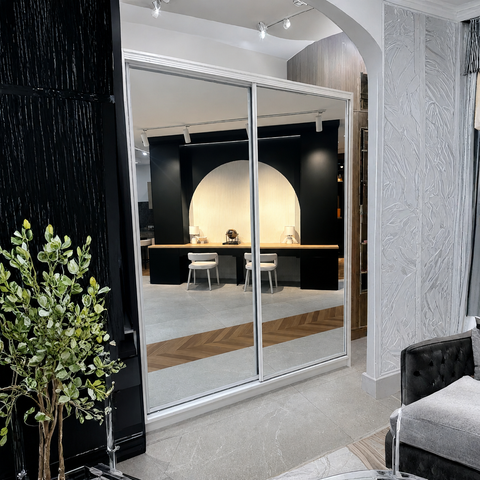 A modern interior space with a large mirrored sliding wardrobe door as the focal product. The reflective doors show a furnished room with a desk, chairs, and arched wall feature, while the actual product is the mirrored wardrobe framed in silver metal.