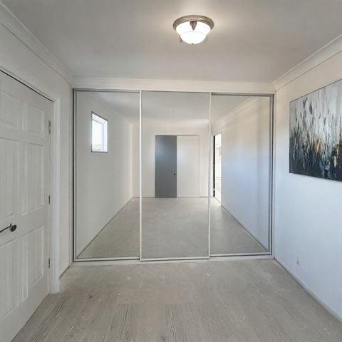 A modern sliding glass door closet or room divider installed in a bright, minimalist room with white walls, light wood flooring, and a ceiling light.