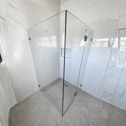 A corner glass shower enclosure with a sliding door and chrome hardware, installed in a modern bathroom with white marble-look walls and gray stone-look floor tiles.